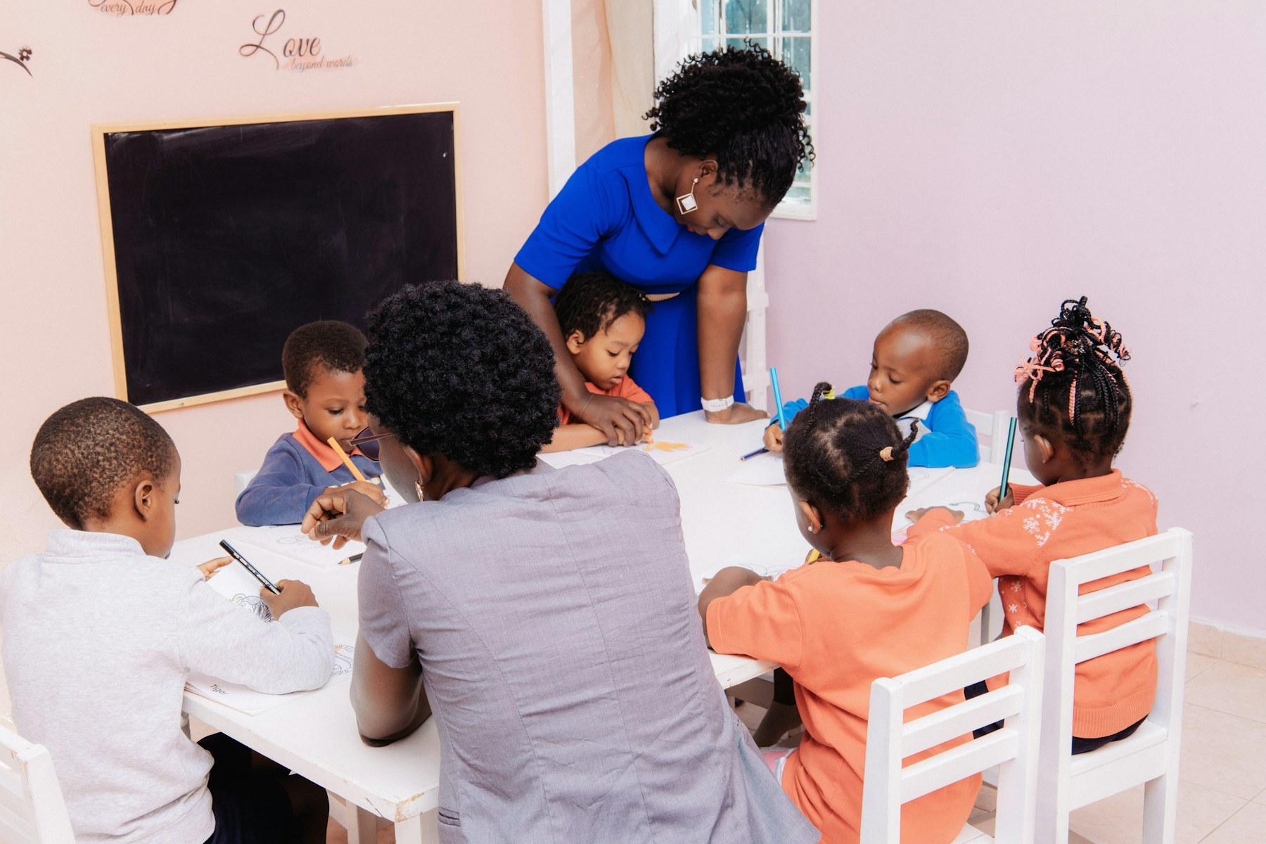 Children draw together at a table with a teacher nearby.
